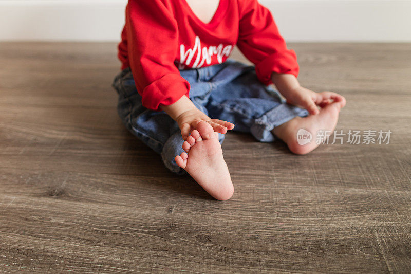 一个可爱的13个月大的圣诞小男孩，光着12个脚趾，穿着复古牛仔裤和红色毛衣，上面写着“妈妈是我的情人”，坐在木质硬木地板上