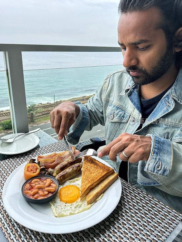 特写画面，印度男子在露天用餐区餐厅餐桌上吃全英式油炸早餐，白色盘子里有早餐，两根香肠，烤豆小蛋糕，培根片，蘑菇，煎蛋，番茄，吐司，重点在前景