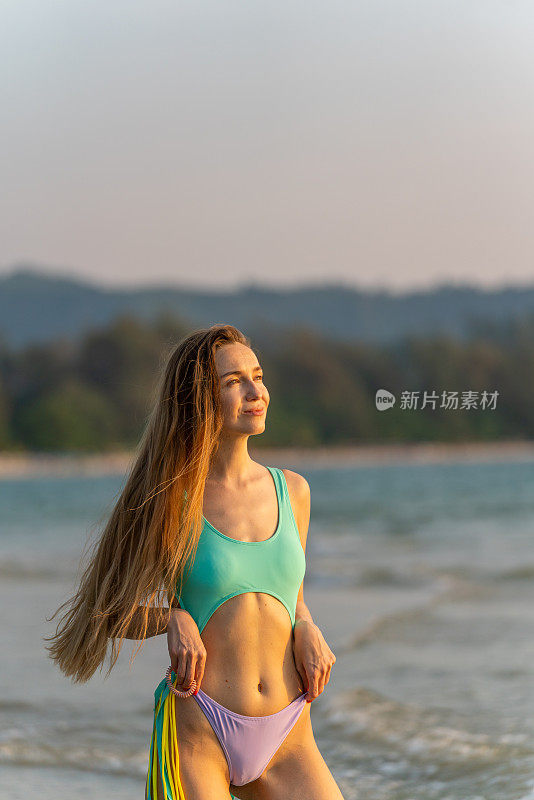 美丽的白人女子在海滩上穿着比基尼，看着大海享受海风和日落的景色