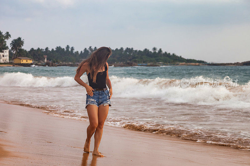 年轻女子在牛仔短裤走在沙滩海浪背景，暑假。旅行者女士漫步热带海岸，放松生活方式。旅游度假概念。复制文本空格