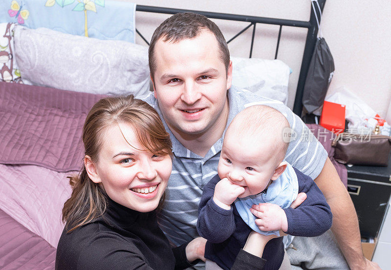 这是一个年轻的白人家庭，一对年轻的男人和女人抱着一个婴儿，他们看着镜头，微笑着。