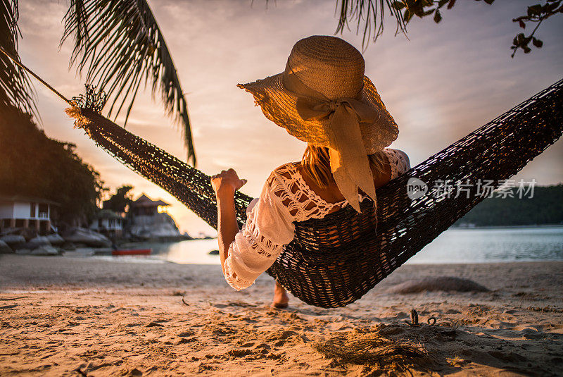 一个女人放松在沙滩上的吊床在日落的后视图。