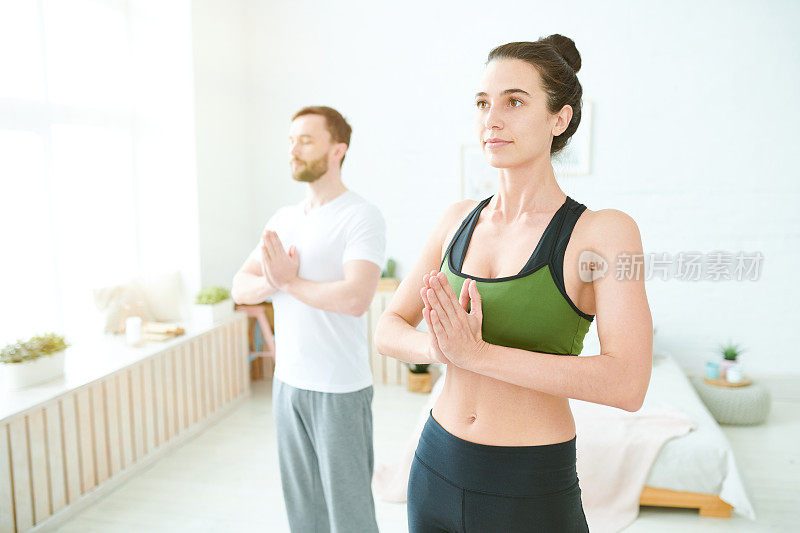 内容平静美丽的年轻女子在运动服装和她的男朋友牵手合十礼，同时准备在家里练习瑜伽