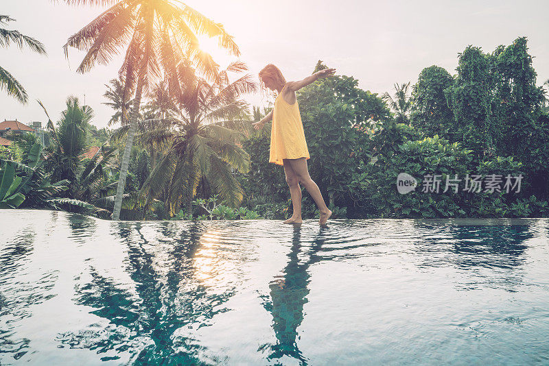 年轻女子走在一个热带天堂目的地的游泳池边缘;女游客在旅游度假池享受度假乐趣的概念