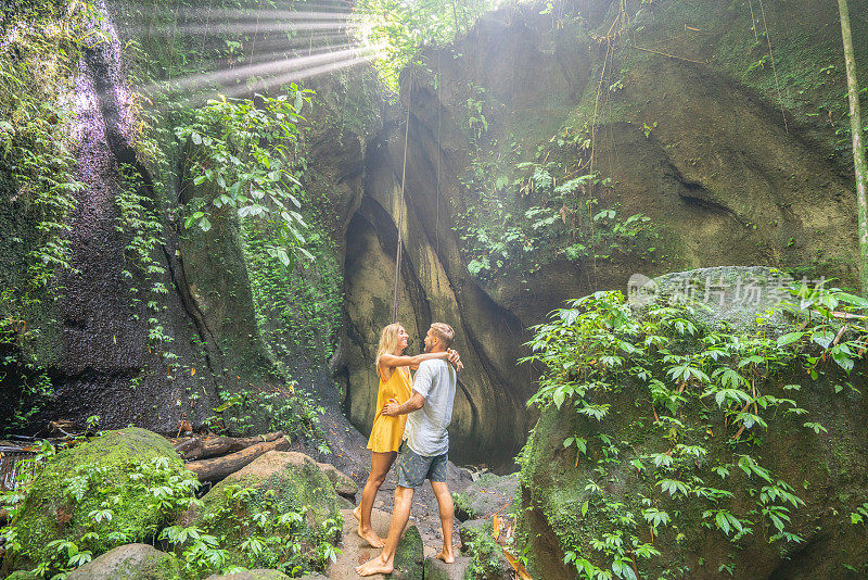 一对年轻夫妇站在热带雨林洞穴里，仰望着从岩石上射下来的壮丽阳光。人们旅行的奇妙欲望自然的概念