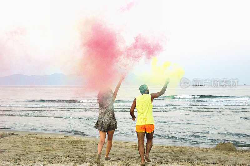年轻的情侣站在沙滩上，手里拿着烟雾弹，浪漫的情侣用黄色和红色的烟雾弹在沙滩上