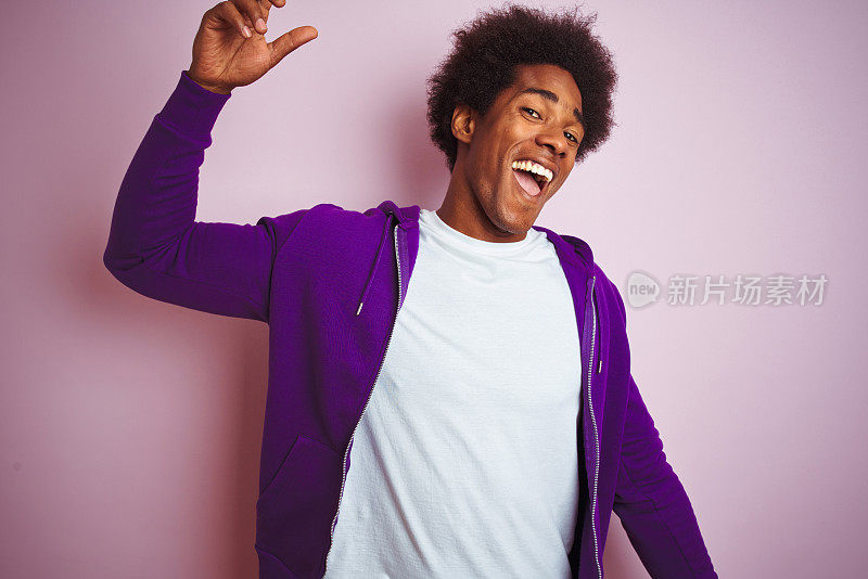 一位年轻的非裔美国人穿着紫色的运动衫站在孤立的粉红色背景上，欢快地跳舞，微笑着，听着音乐，轻松而自信