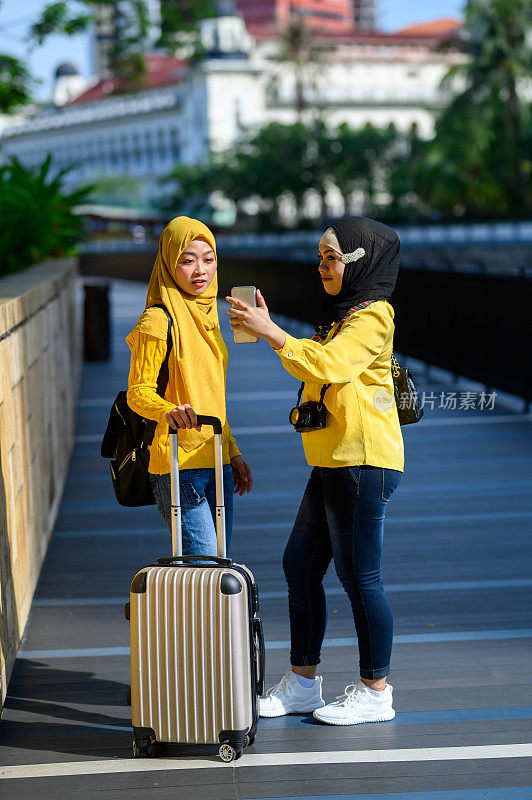 两名女性穆斯林旅行者戴着头巾，用智能手机自拍在城市街道上