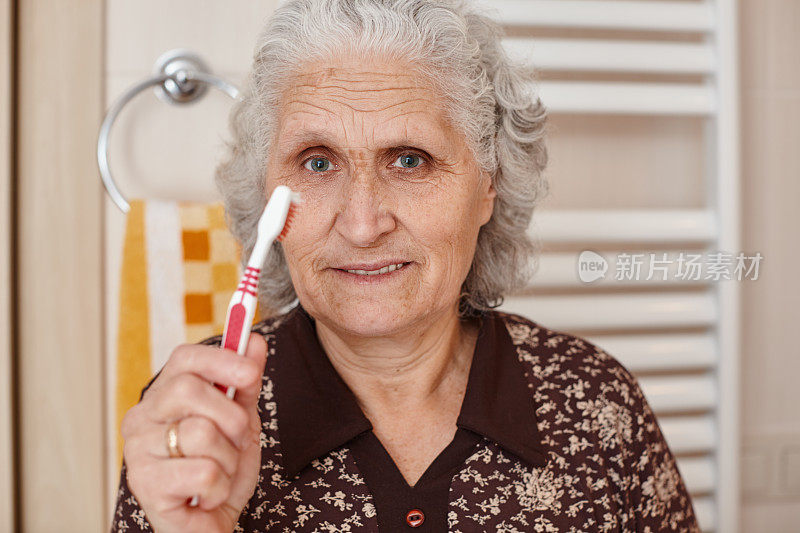 美丽的老妇人与灰色的头发拿着一个牙刷在手里，而看着镜头在浴室的镜子和水槽