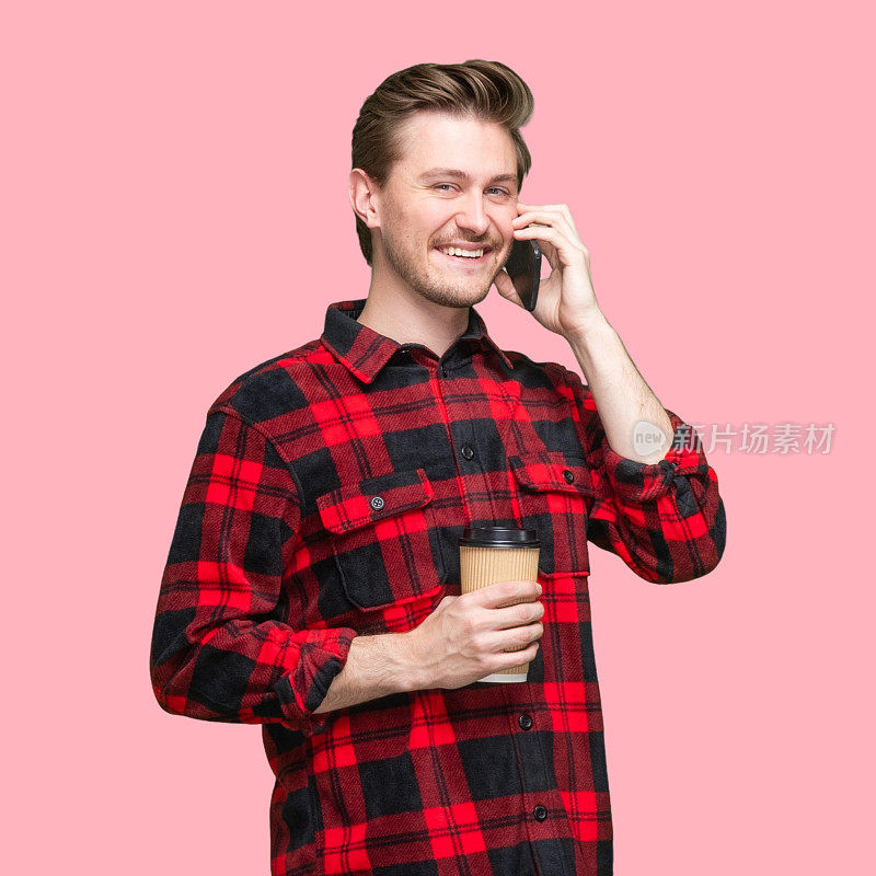 白人男性站在有色背景前，穿着格子衬衫，拿着咖啡杯，使用智能手机