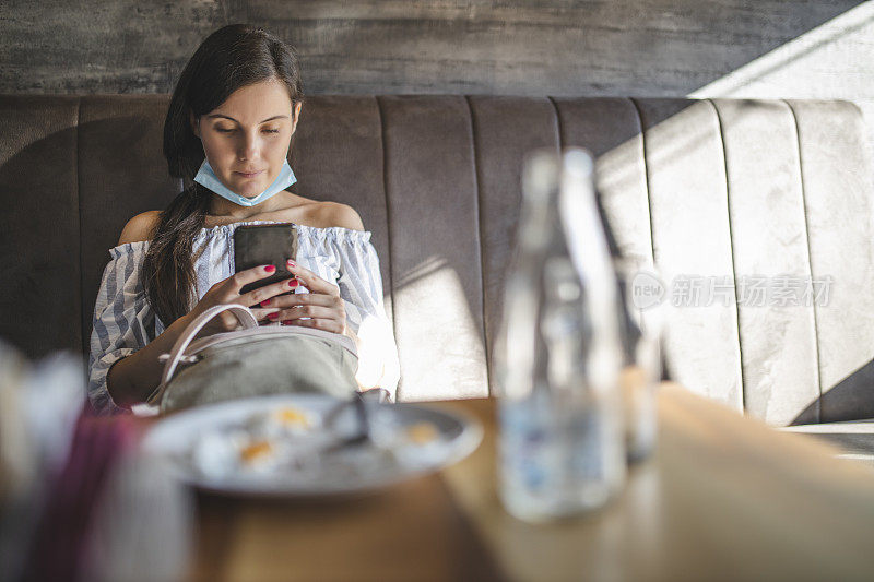 年轻女子戴着外科口罩，在疫情期间使用手机，在咖啡馆消磨时间