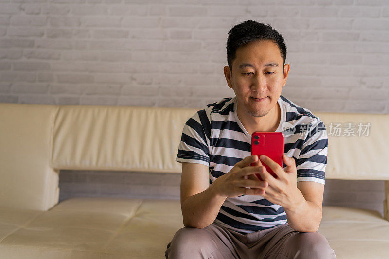 亚洲男人手里拿着智能手机，家里有复印空间，坐在沙发上