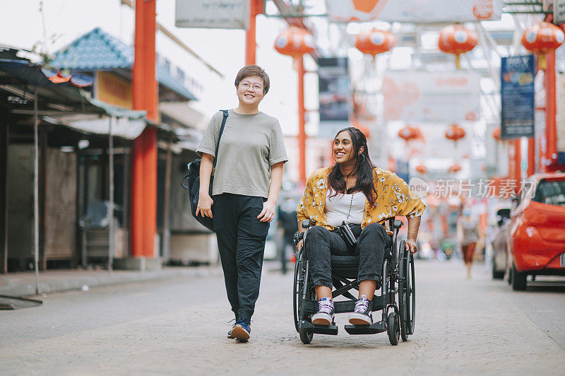 亚洲华人中年妇女和她的印度残疾女性朋友肩并肩走在城市街道上的轮椅上