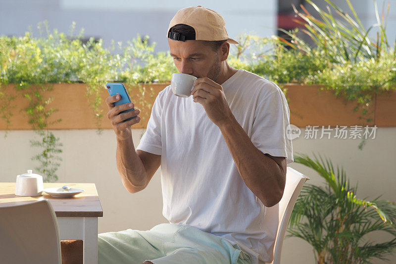 男人坐在夏日的咖啡馆在阳光明媚的早晨喝咖啡，看着蓝色的手机。生活方式的人。
