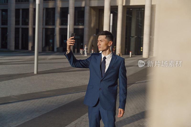 一位身穿蓝色西装的年轻亚洲经理站在商务中心前的广场上自拍