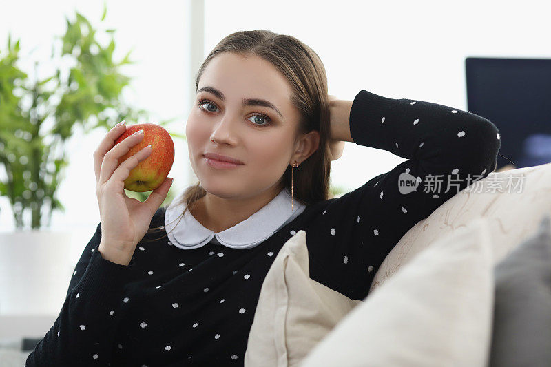 漂亮的年轻女子舒舒服服地坐在家里的沙发上，吃着新鲜的苹果果