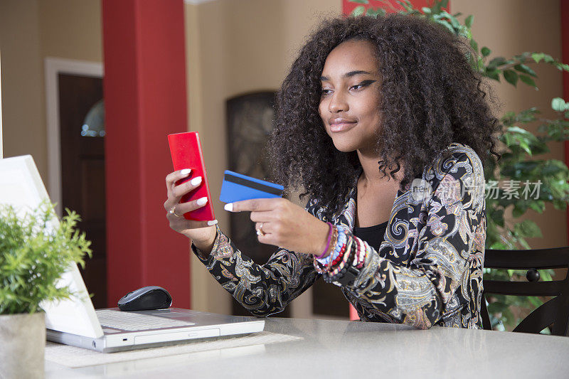 可爱的非裔美国青少年在网上购物。她坐在厨房吧台旁。笔记本电脑、智能手机和信用卡。