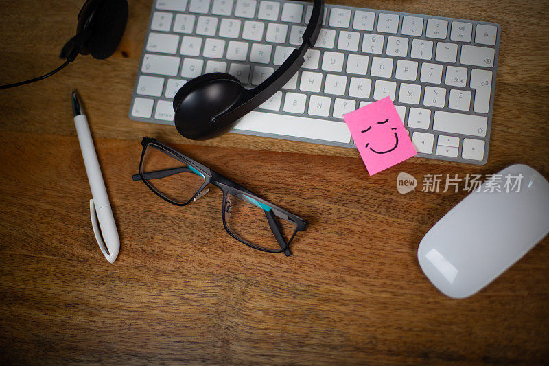 工作中幸福和幸福的象征。配备电脑、眼镜和耳机麦克风的工作站。键盘上贴着一张带微笑的粉色便签。