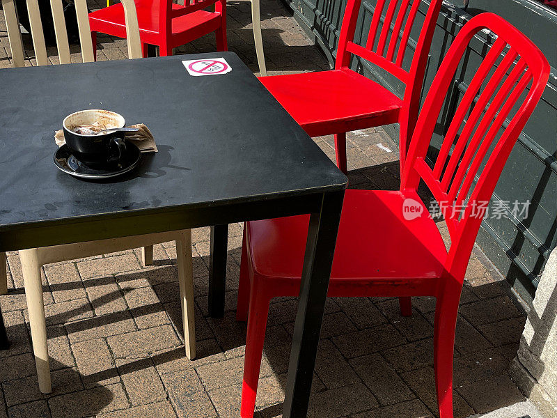 路边餐厅外的方桌和红色椅子的特写图像，人行道上的咖啡馆露天餐厅，编织单色空咖啡杯和茶托，没有吸烟的标志，阳光明媚的日子，重点在前景