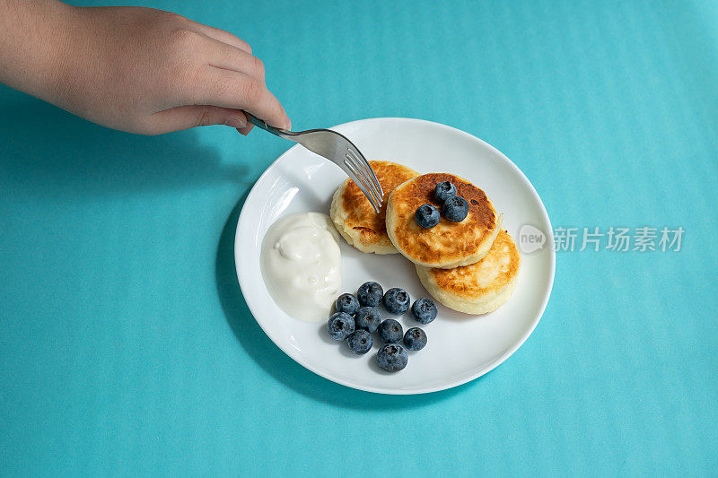 手握叉子和刺煎饼。芝士蛋糕放在白色盘子里，配酸奶油和蓝莓