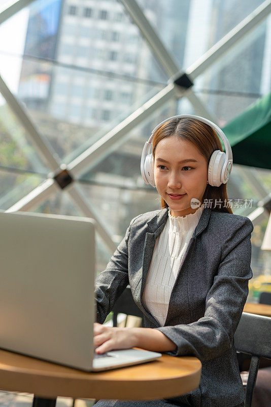 混合式工作风格。年轻的亚洲女性成年人与笔记本电脑在咖啡馆工作。戴白色耳机的女商人
