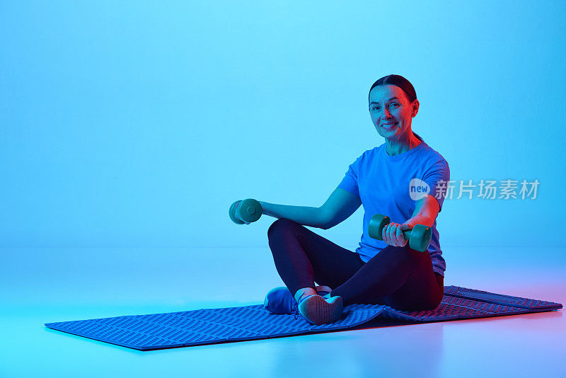 微笑，积极，成熟的女人坐在健身matt和训练哑铃对蓝色工作室背景霓虹灯