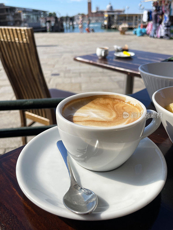 白色杯子和碟子卡布奇诺咖啡与不锈钢茶匙在露天餐桌上的特写图像，威尼斯泻湖滨水的背景，重点在前景