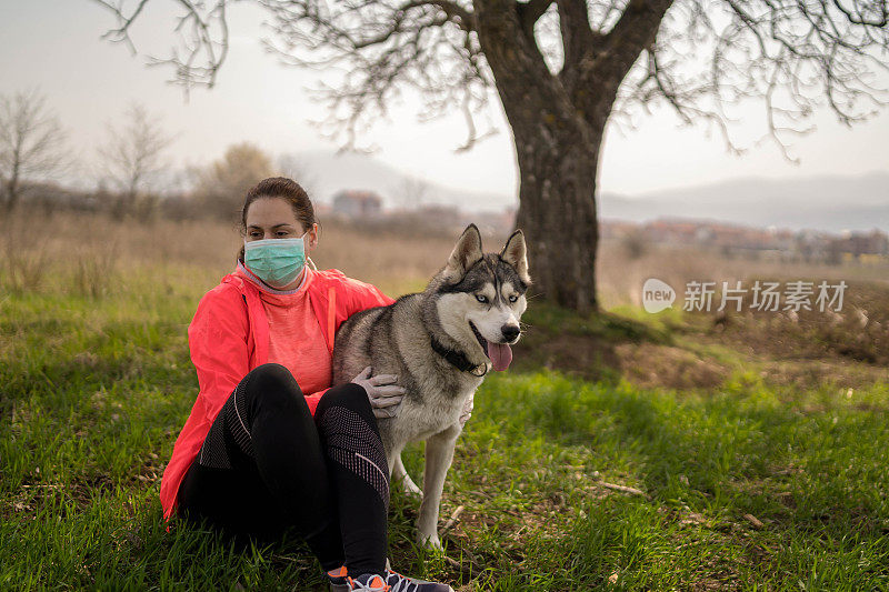 戴着防护口罩的年轻美女和她的白狗在公园外面的草地上休息