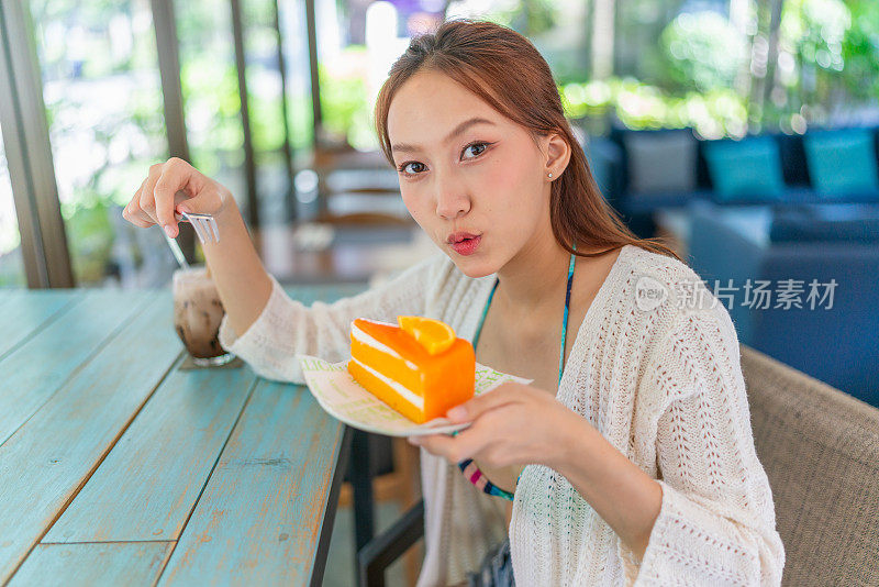 年轻的亚洲女子在海滩咖啡馆喝着冰拿铁，一边享用着一片橘子蛋糕。穿着沙滩装的女性数字游牧民在度假氛围中休息。穿着沙滩装的亚洲女人在咖啡馆里放松。