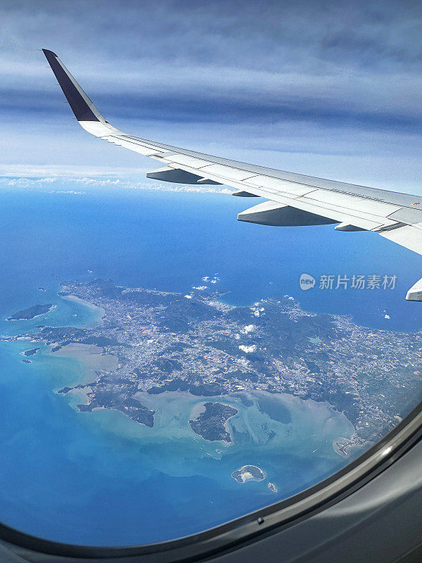 从飞机窗口看到的图像，飞机机翼在云层下，海洋周围的陆地地形，小翼和板条，蓝天，重点在前景