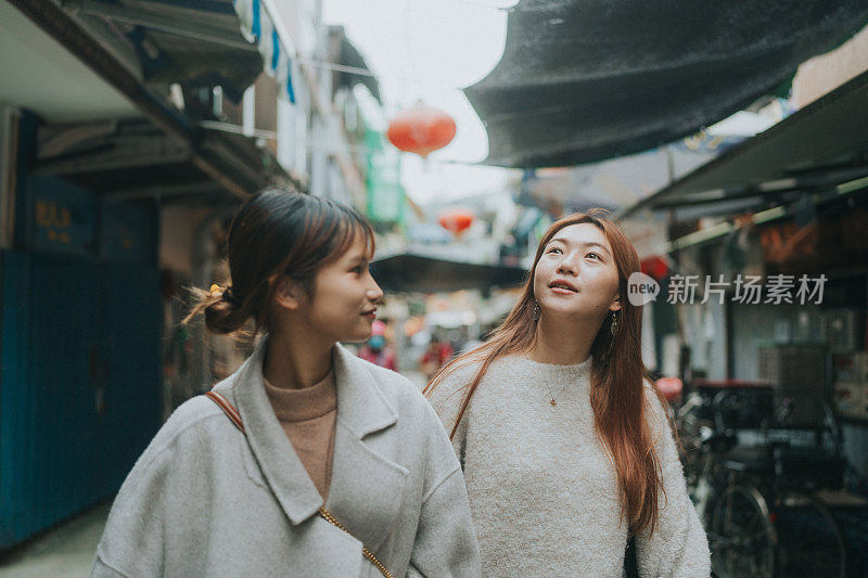 在香港长洲，两名亚洲女子漫步在商店和自行车林立的街道上，悠闲地漫步，可能是在探索或观光