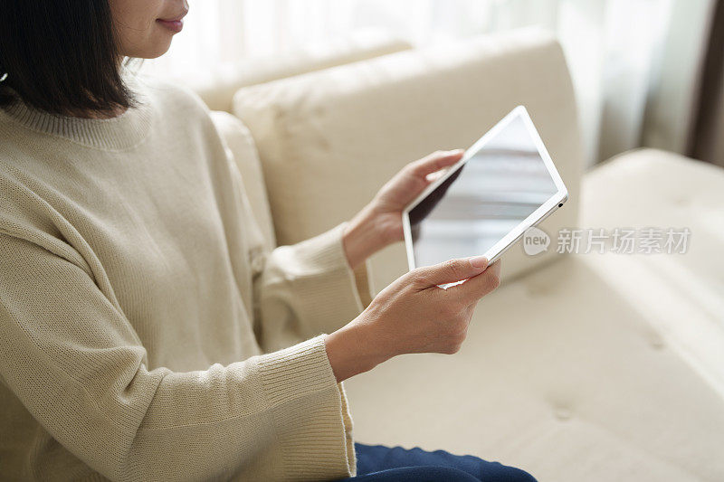 在特写镜头中，一位亚洲女性坐在沙发上，她的手正在使用电子平板电脑，同时在家工作，阳光温柔地照在她身上。