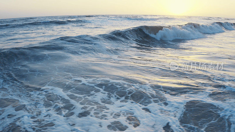 在黄金时段飞越海洋。巨大的海浪在海面上泛起泡沫，溅起水花。电影式的无人机拍摄，落日波浪形的海景。海洋和天空背景
