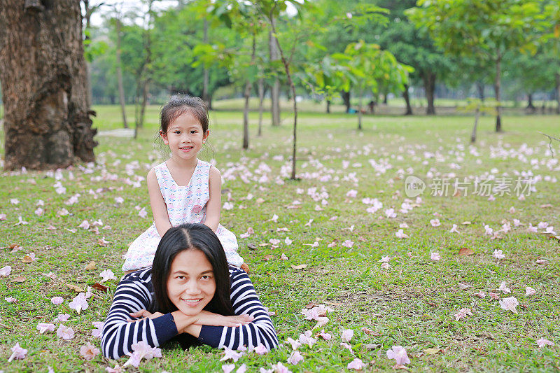 欢快的小女孩骑在妈妈的背上，躺在绿色的田野上，满是粉红色的花朵。幸福美满的家庭。