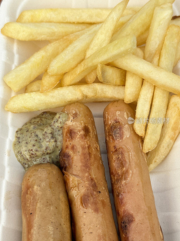 矩形的全画幅图像，一次性托盘包含三个德国法兰克福香肠，部分薯条与一团芥末，高架视图