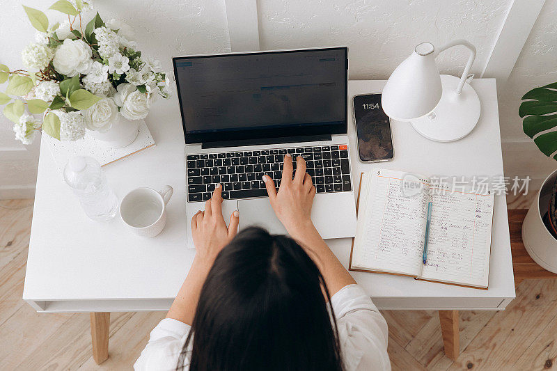 一个女人在白色的办公桌上用笔记本电脑打字，桌子上有台灯、笔记本、一瓶饮用水和花瓶里的花。自由职业的概念，远程工作，在线学习。在家工作
