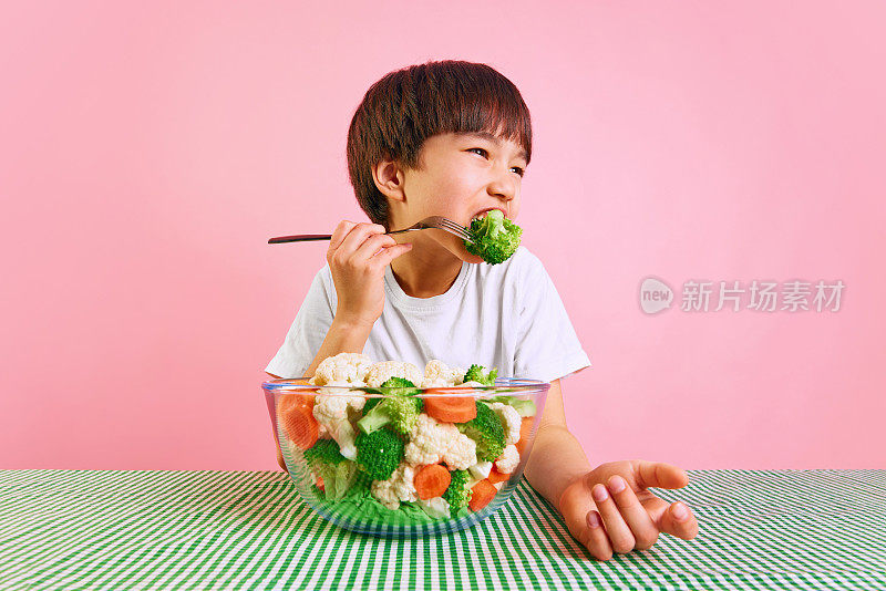 小男孩，孩子坐在桌子旁吃着花椰菜，碗里盛满了粉红色背景的新鲜蔬菜