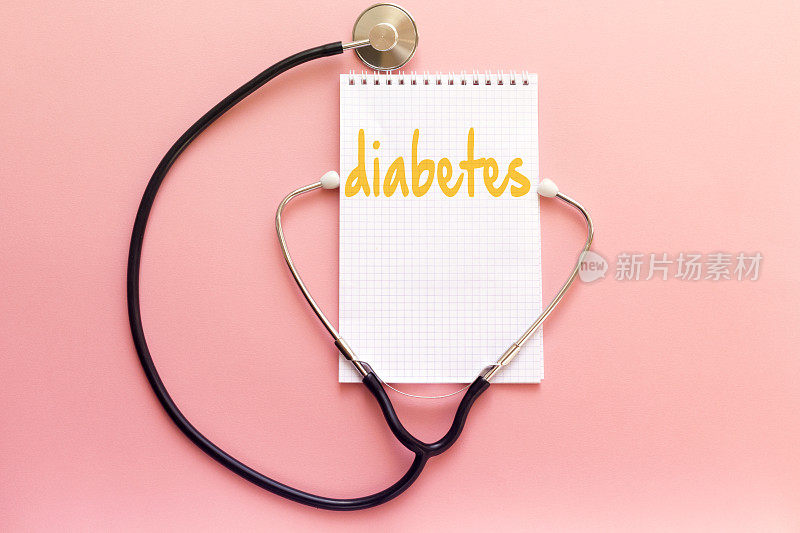 疾病糖尿病概念书写在白色笔记本上，听诊器在粉红色背景上