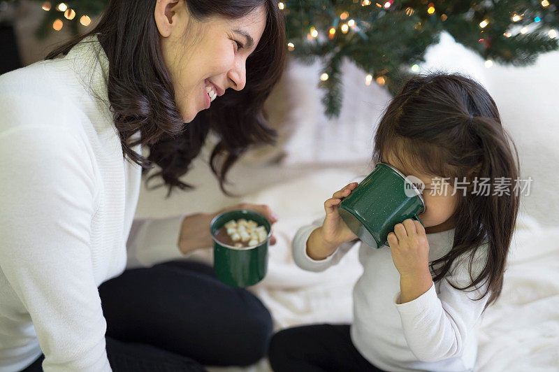 年轻的民族女孩和她的妈妈一起在圣诞树旁喝热巧克力