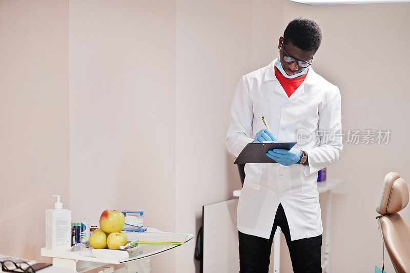 戴眼镜和面具的非裔美国男医生在诊所里摆姿势，手里拿着黑色的写字板，写着什么。