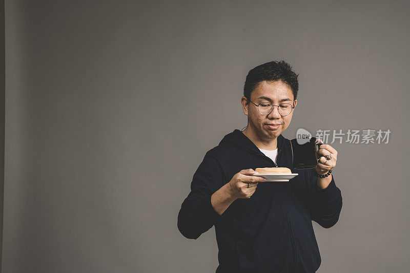 一个亚裔华人男子拿着一杯咖啡和一盘灰色背景的甜甜圈