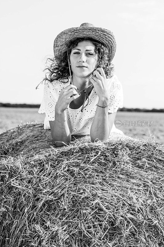 可爱的年轻女子坐在一个干草堆上，背景是一张麦田的照片