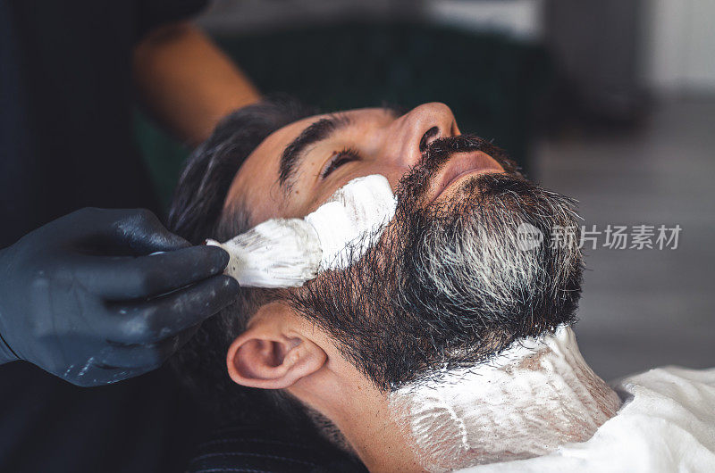理发师戴着乳胶手套，用刷子将膏霜给顾客勾勒出胡须轮廓
