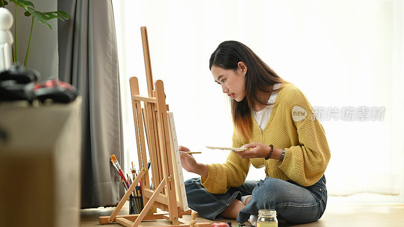 一位年轻女艺术家坐在她的工作室的地板上在画布上作画的侧视图。