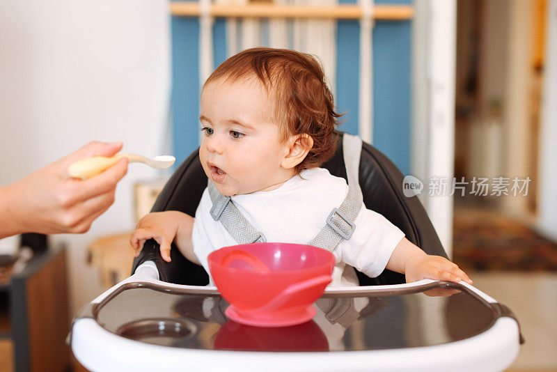 微笑的8个月大男婴坐在家里的高椅子上，妈妈用勺子给他喂固体食物