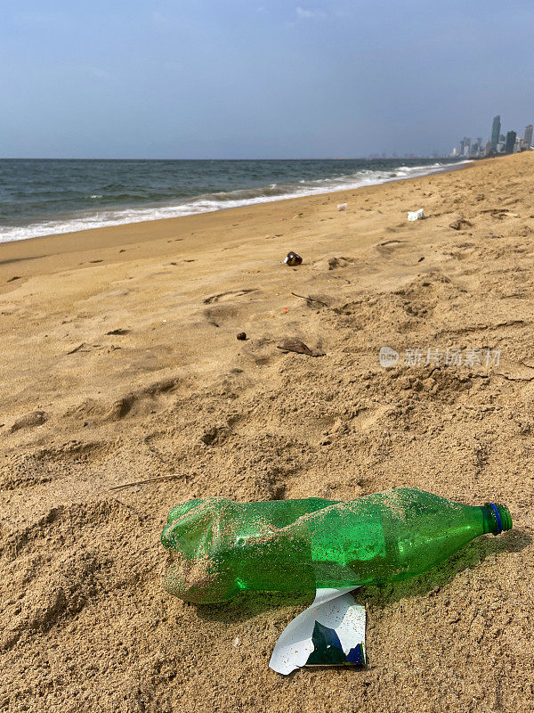 特写图片:退潮时被冲到沙滩上的破碎的绿色塑料水瓶，海岸上的海洋垃圾和污染，乱扔的沙子，肮脏的海滩，科伦坡市摩天大楼的海岸线视图，拉维尼亚山海滩