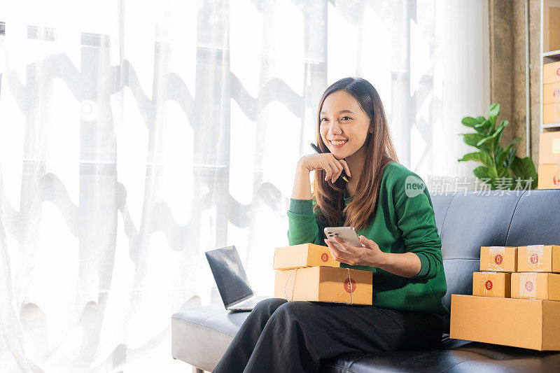亚洲女性小企业主的概念，拿着智能手机，高兴地接收客户的订单信息，在家里用笔记本电脑网上购物，准备发送包裹