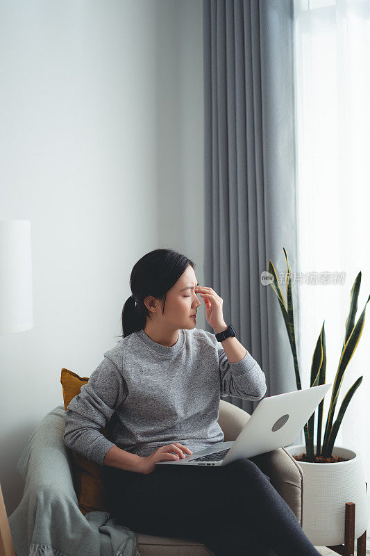 一名亚洲女性坐在家里的客厅里使用笔记本电脑，饱受眼睛疼痛之苦。