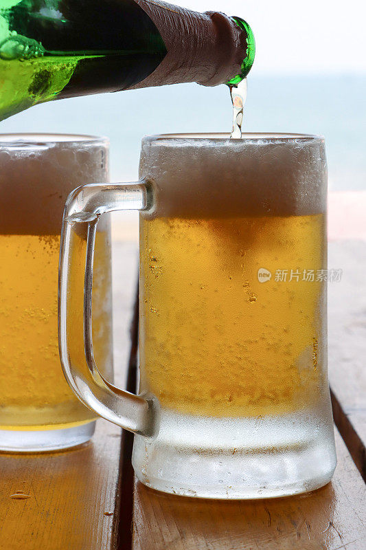 啤酒被从绿色啤酒瓶倒进玻璃酒杯的图像，在凝结与气泡形成泡沫的泡沫顶部的啤酒，品脱玻璃杯在海滩酒吧户外桌子上，关注前景