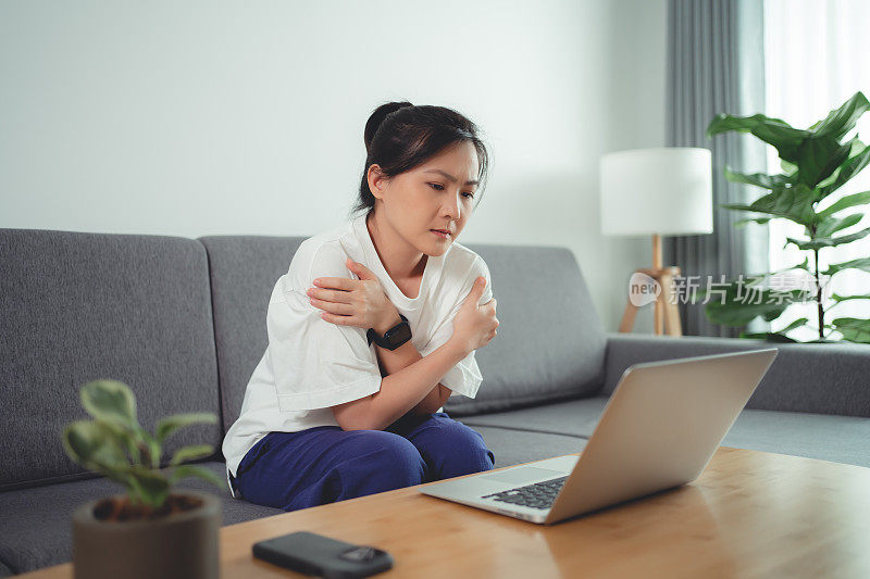 一名亚洲妇女坐在家里的沙发上，用笔记本电脑工作，发烧了。